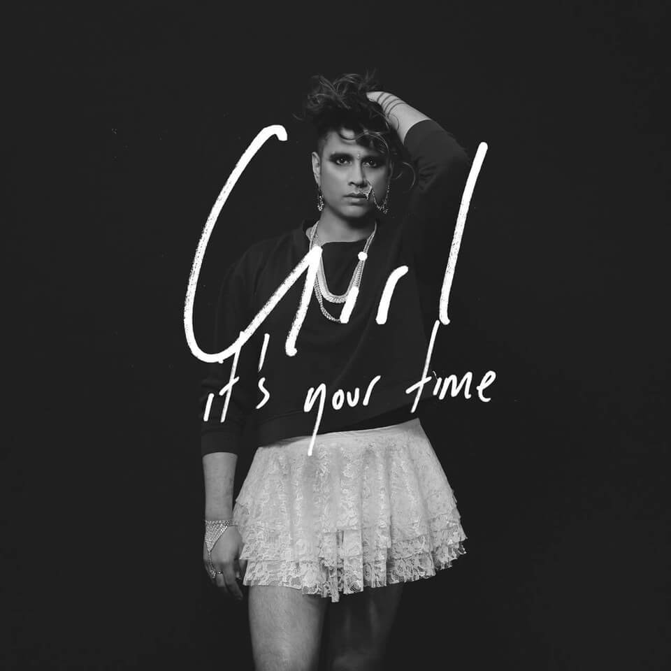 A B&W photo of a brown-skinned femme in her mid-30s standing in front of a black background wearing a plain black sweatshirt and lacy white skirt, overlaid with white handwritten text, “Girl It’s Your Time”.